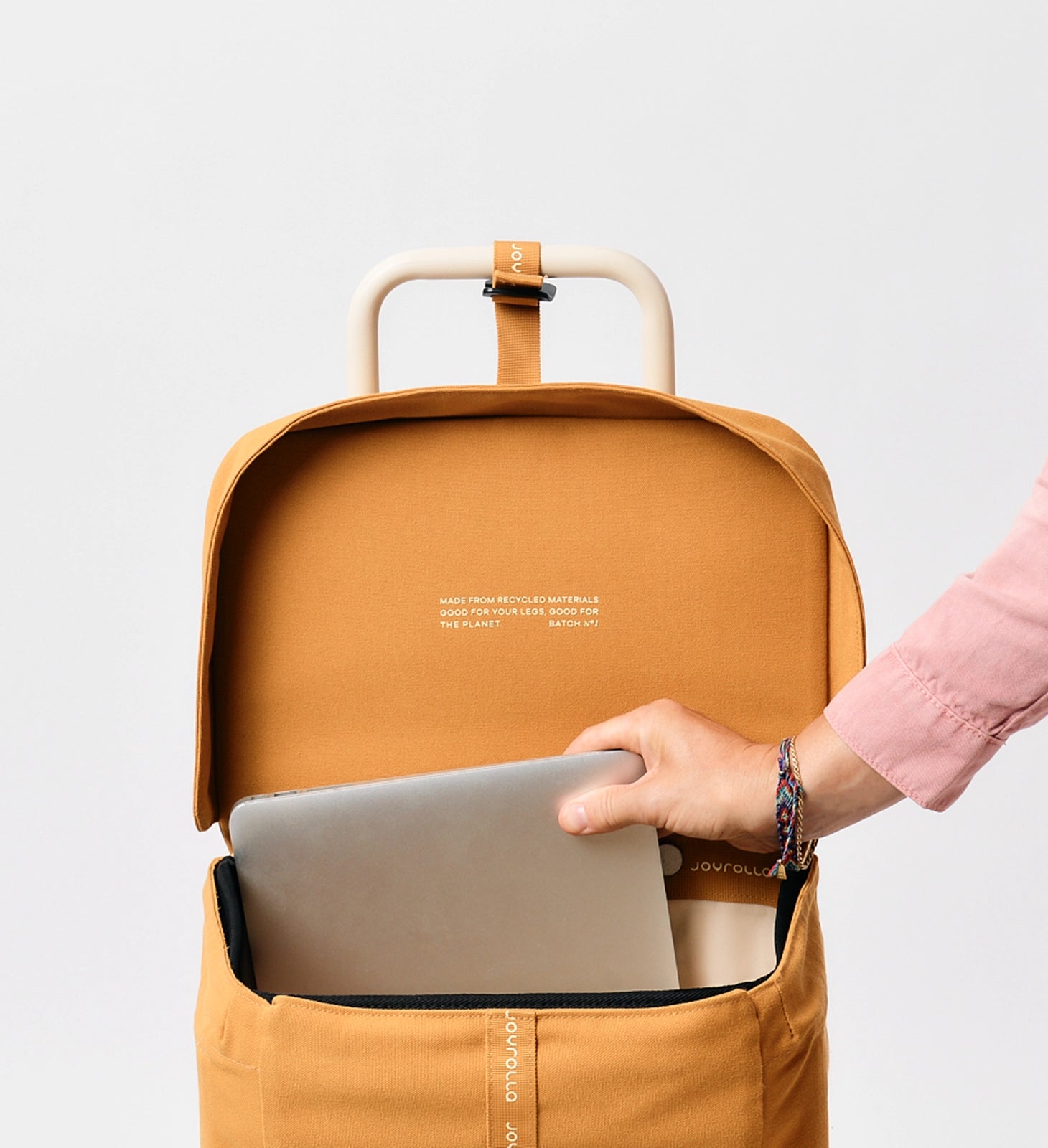 A female hand placing a laptop into the ochre Joyrolla Cart bag, with the lid lifted up for easy access. The image captures a close-up view of the hand holding a laptop and gently sliding it into the open bag. The vibrant ochre colour of the Joyrolla Cart bag stands out, adding a touch of style to the scene. This image demonstrates the versatility and functionality of the Joyrolla Cart, making it suitable for various uses, including carrying electronics securely.